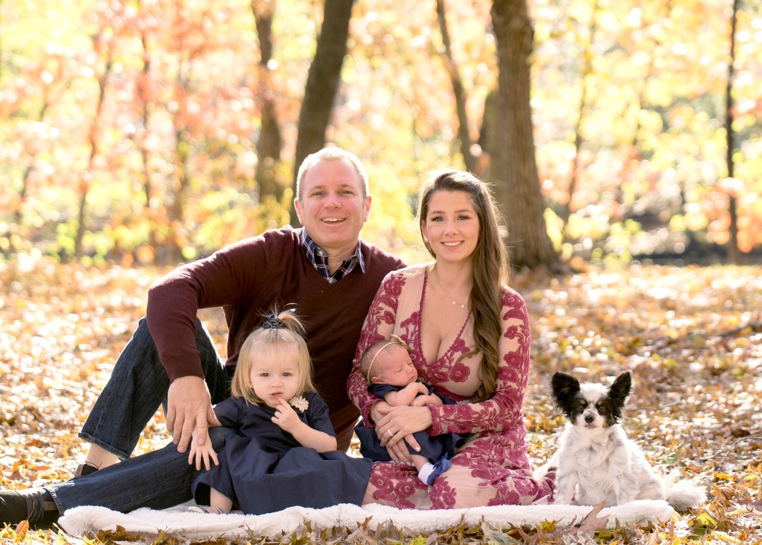 Family portrait with parents, children and small dog sitting on blanket surrounded by autumn foliage.