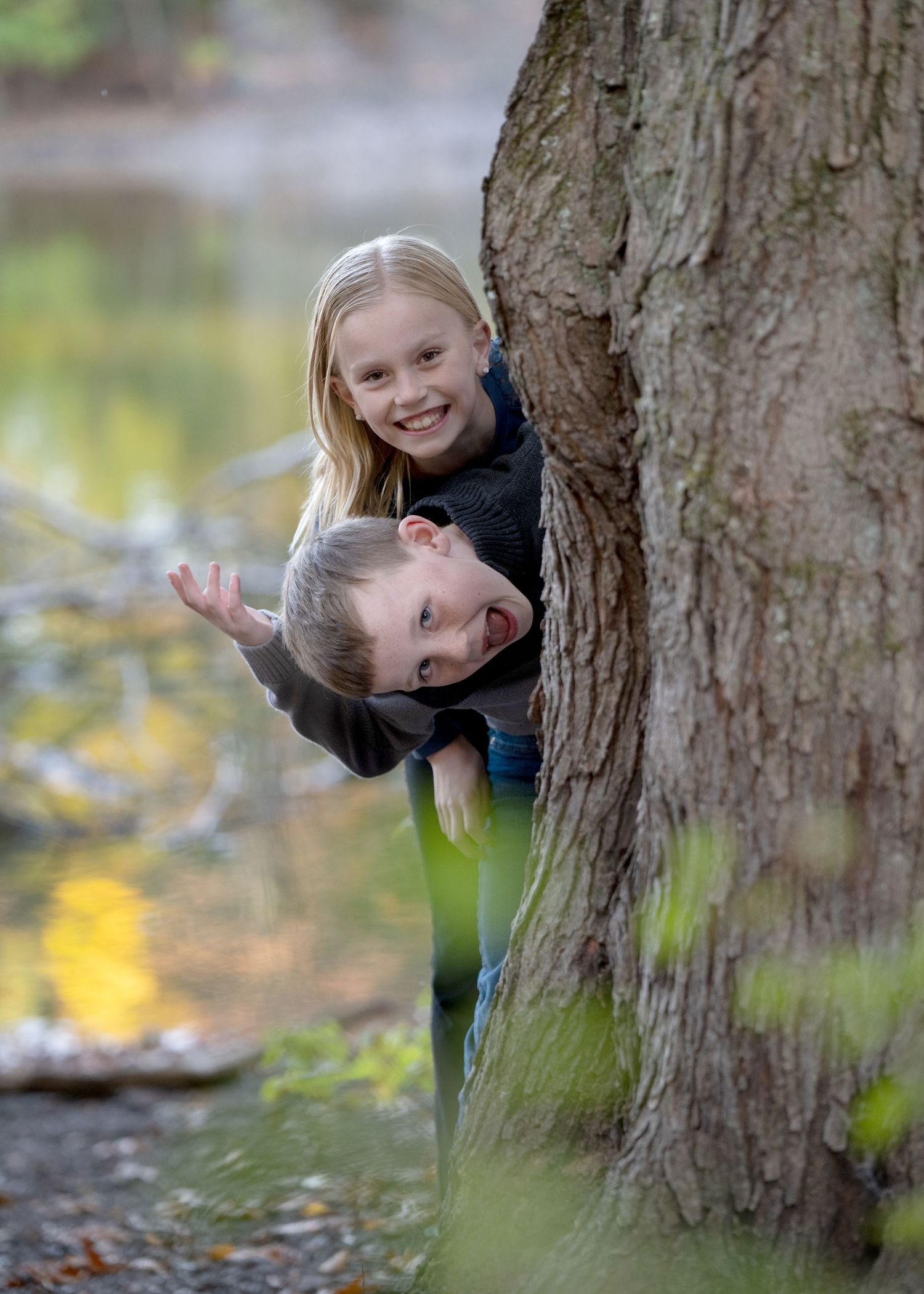 Two children playfully peek out from behind a large tree trunk in a park setting.