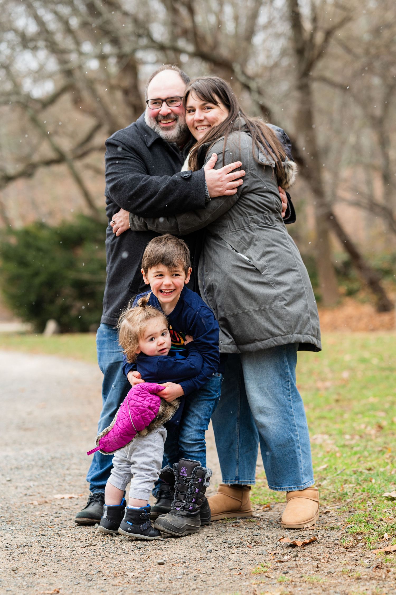 Family of four hugging outdoors in winter, smiling together on a park path surrounded by bare trees.