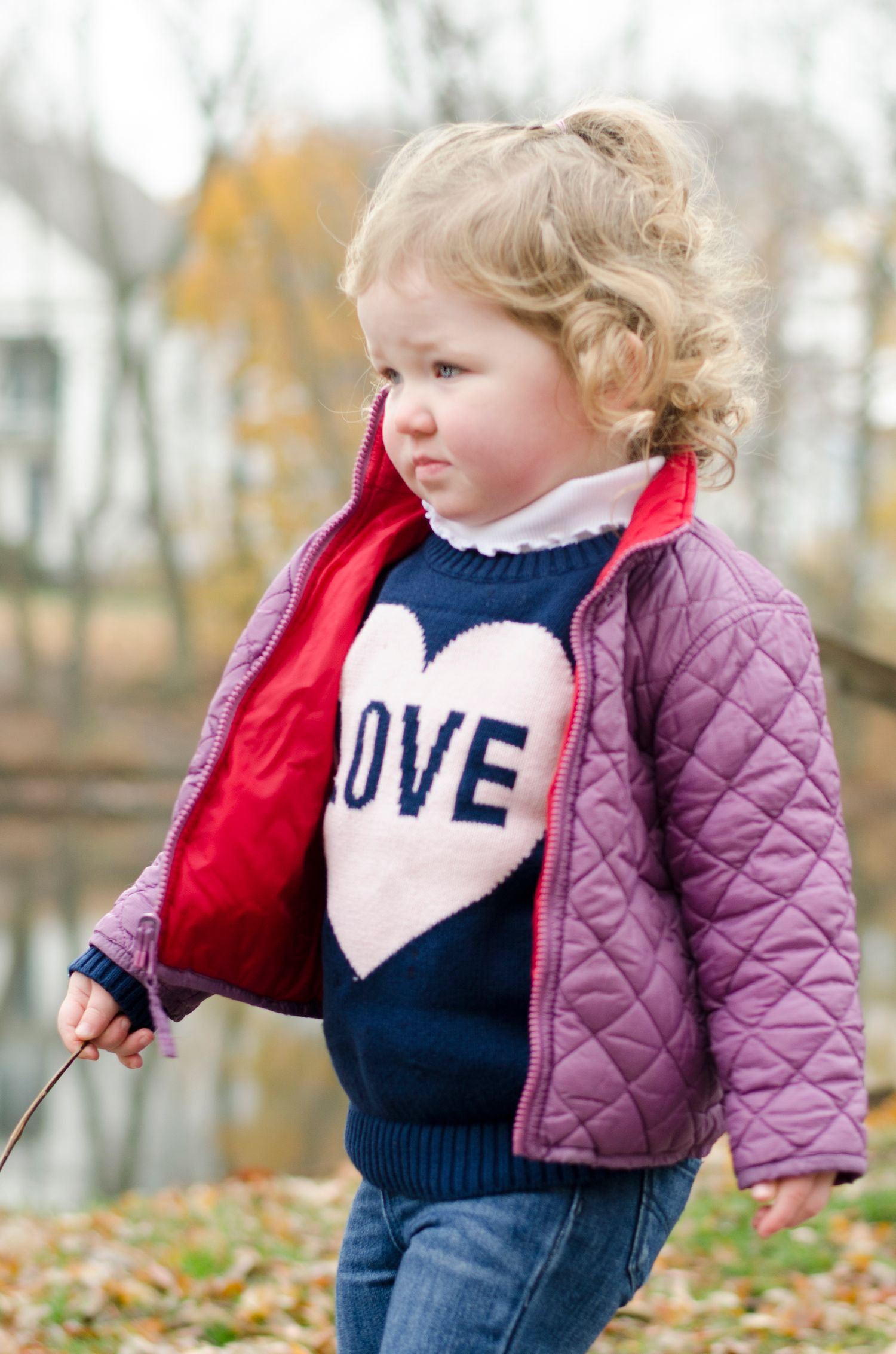 Young child wearing a navy sweater with LOVE text and purple quilted jacket stands in autumn foliage.