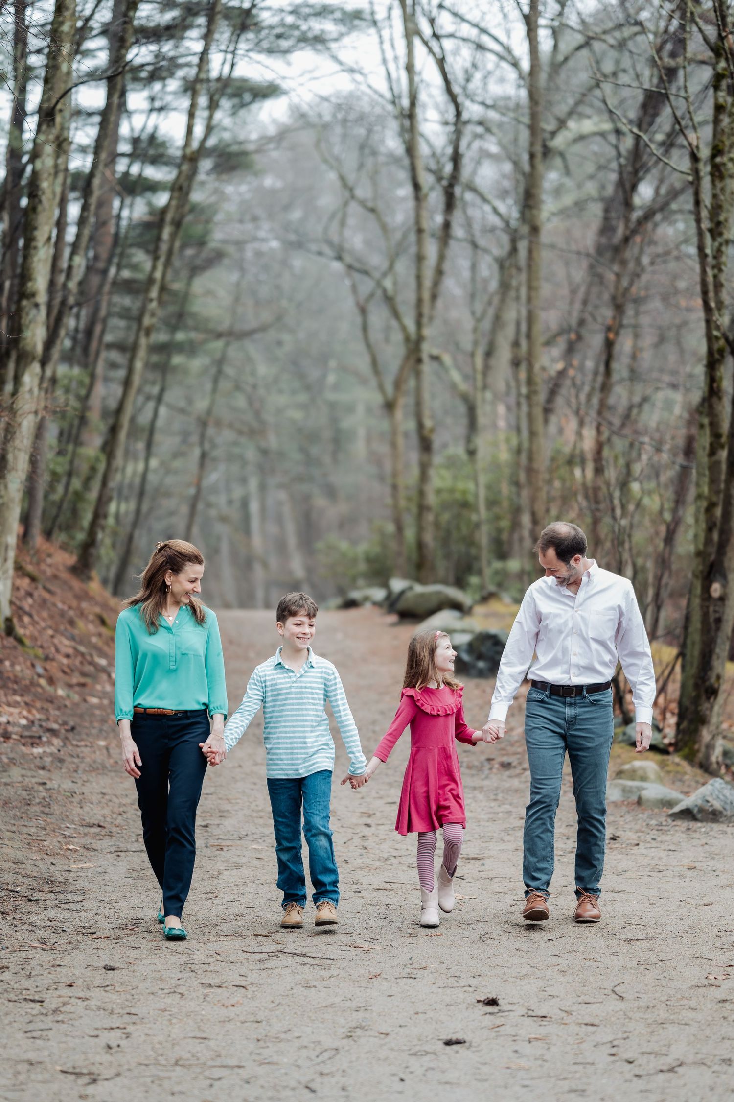 Family of four holding hands walking on a forest path, parents and two children smiling outdoors.