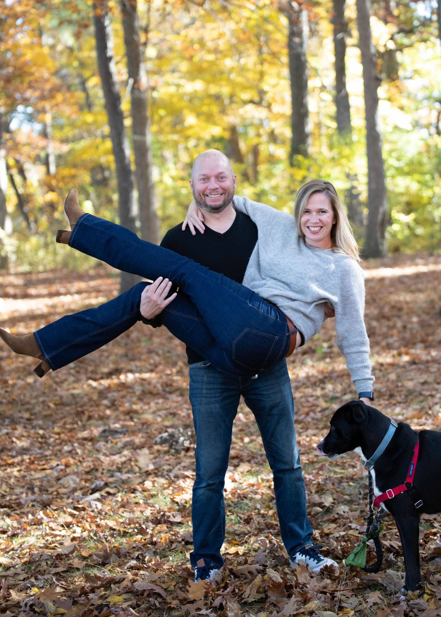 Playful couple posing in fall forest with their black dog as man carries woman horizontally.