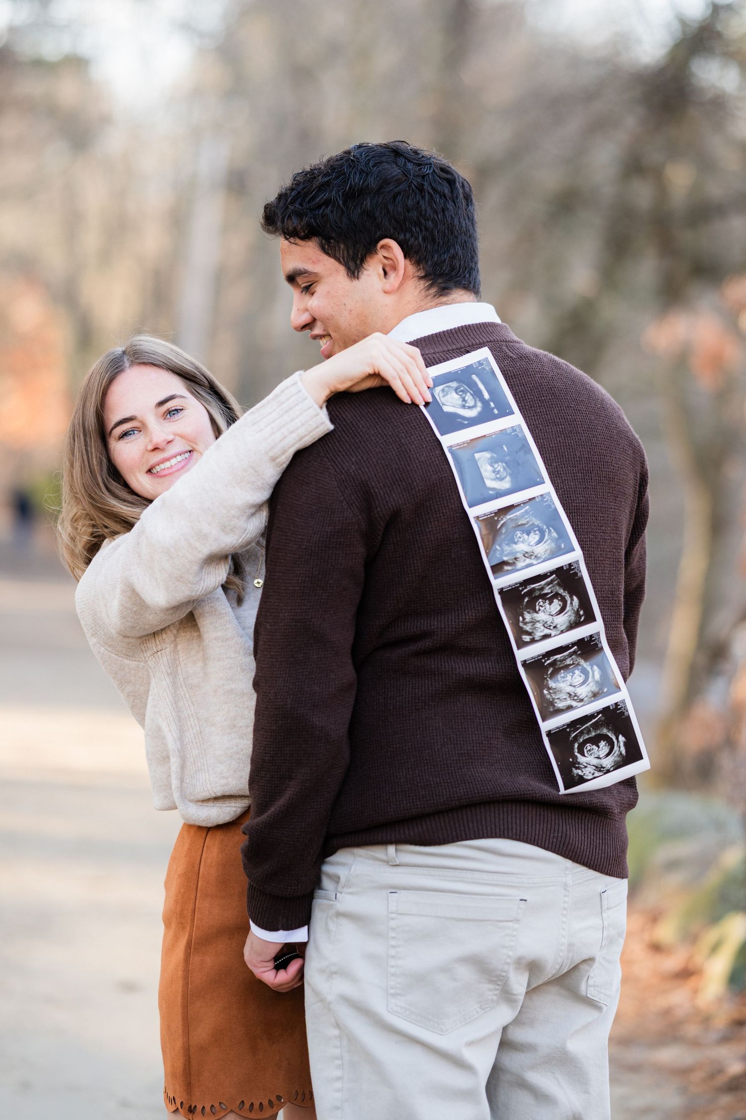 Couple sharing a pregnancy announcement outdoors, man showing ultrasound photos on his back while woman smiles warmly.
