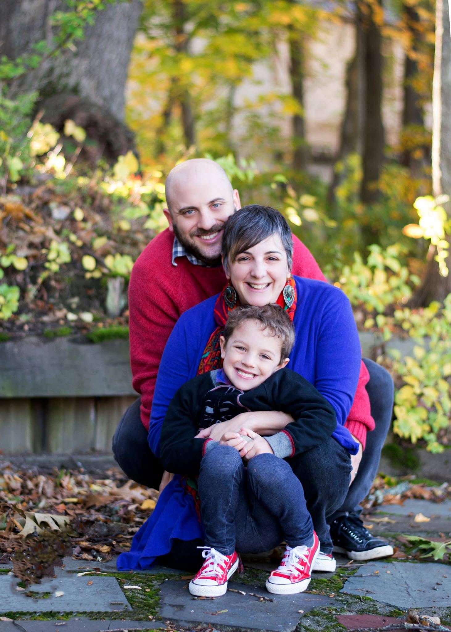 Family portrait outdoors on stone wall with autumn trees in background.