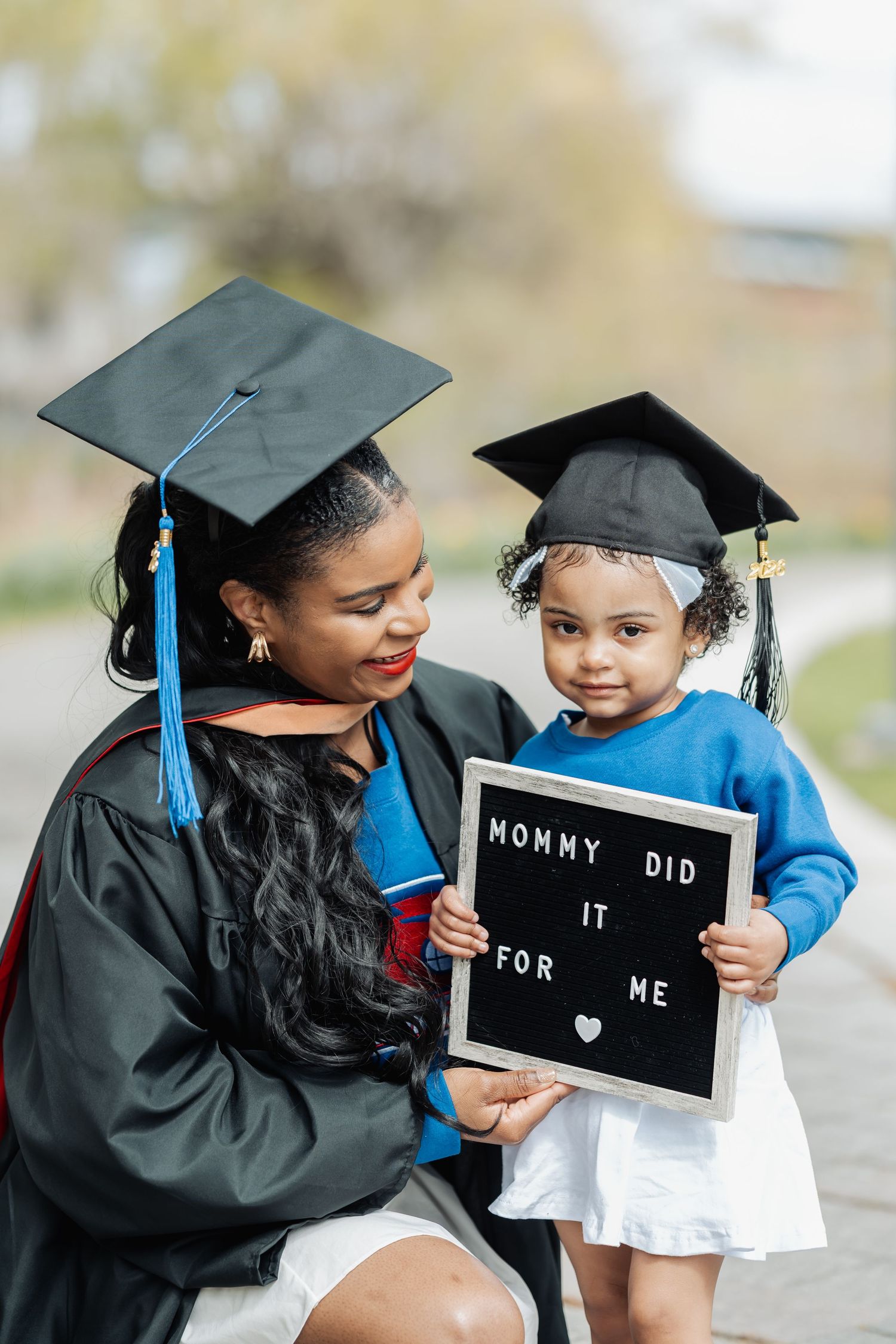 A proud mother in graduation gown and cap poses with her toddler holding a sign reading Mommy Did It For Me.