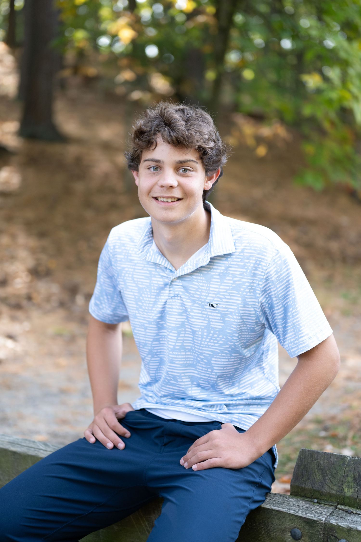 Smiling teenage boy in a light blue polo shirt sitting on a wooden bench outdoors in a wooded park setting.