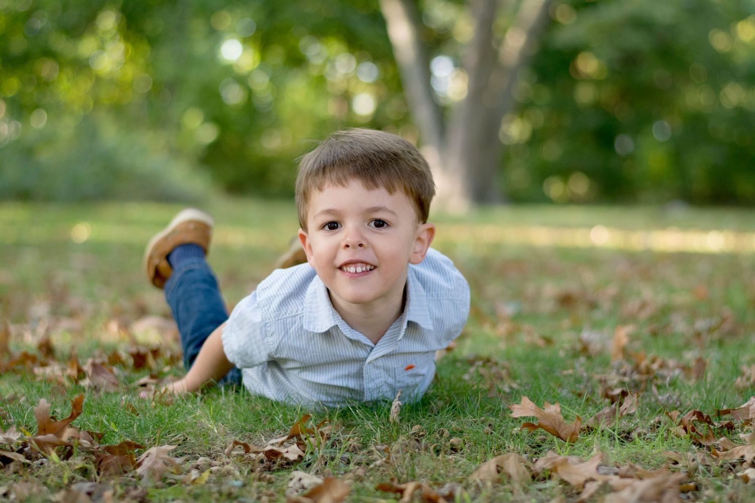 Child lying on ground smiling at camera with autumn leaves scattered around.