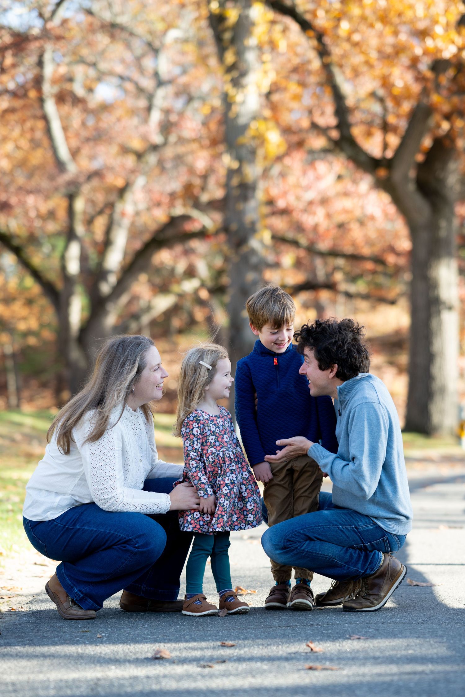 Family of four enjoying autumn park, parents crouching to chat with young children on a tree-lined path.