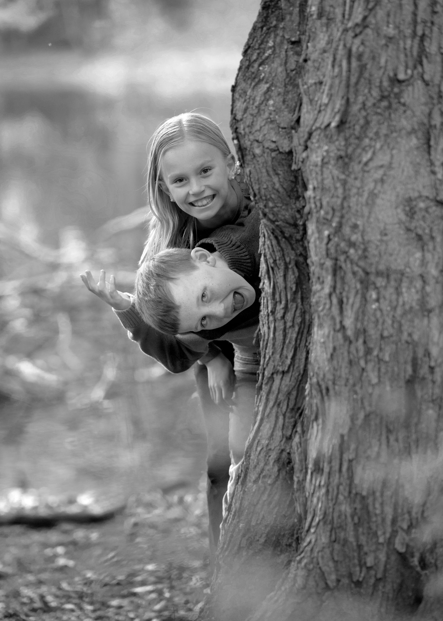 Black and white photo of children playfully peeking around large tree trunk.