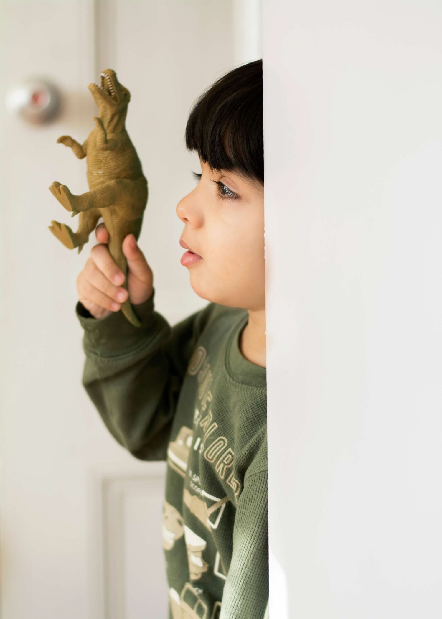 Child in green sweatshirt playing with toy dinosaur figure against white wall.