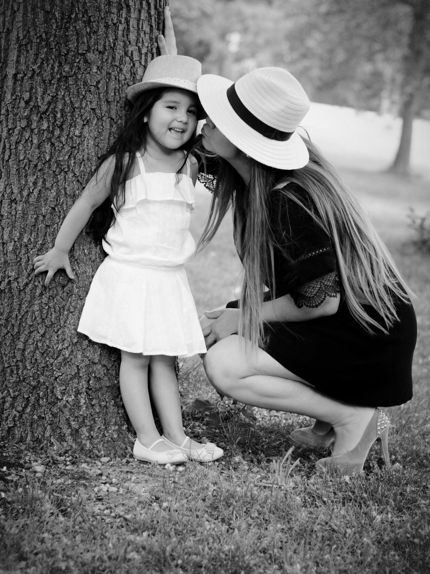 Black and white photo of a parent and child sharing a tender moment by a tree trunk outdoors on a summer day.