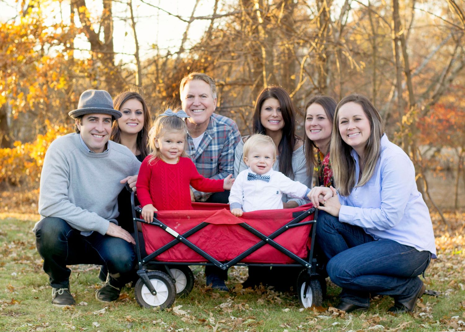 A group sits together with small children in a red wagon during an autumn outdoor photo session.