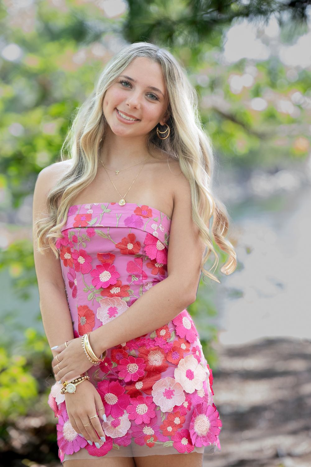 Young woman in pink floral strapless mini dress with gold jewelry, smiling outdoors near greenery.