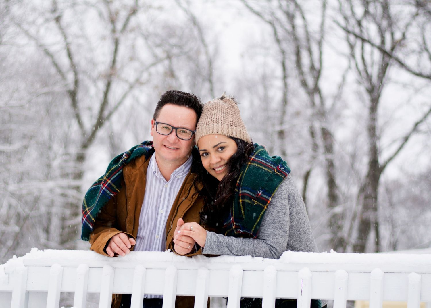 A couple leans on a white railing surrounded by a snowy winter landscape.