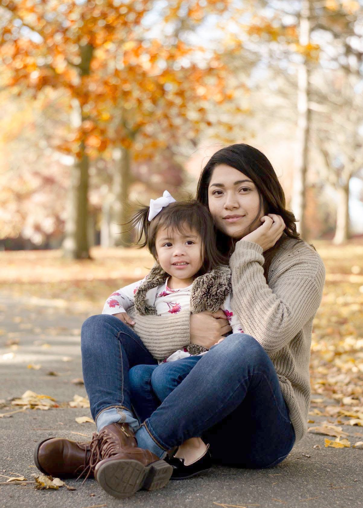 Parent and child sitting together on ground with fall foliage in background.