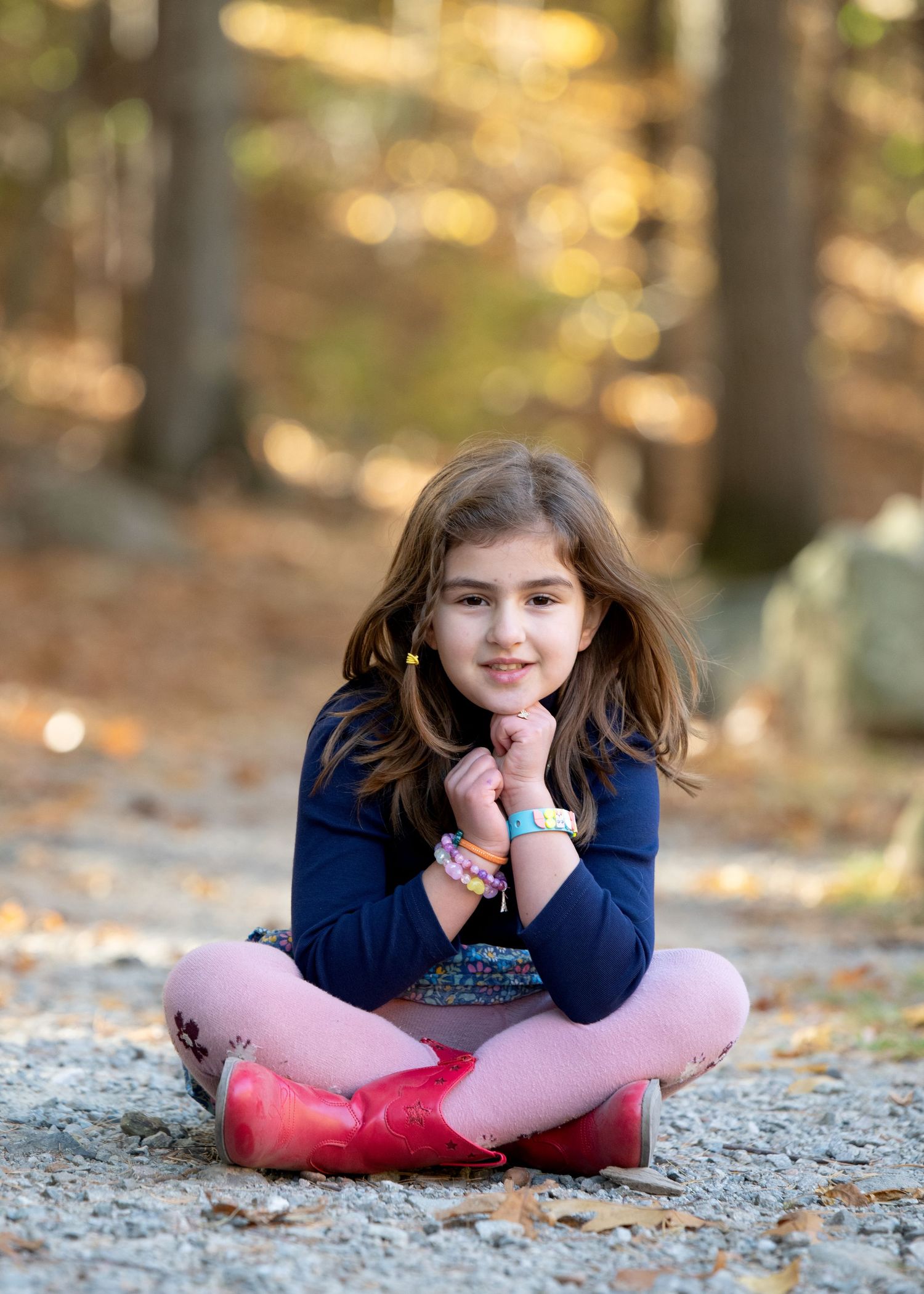 A child in pink boots sits cross-legged on a rock in an autumn forest setting.