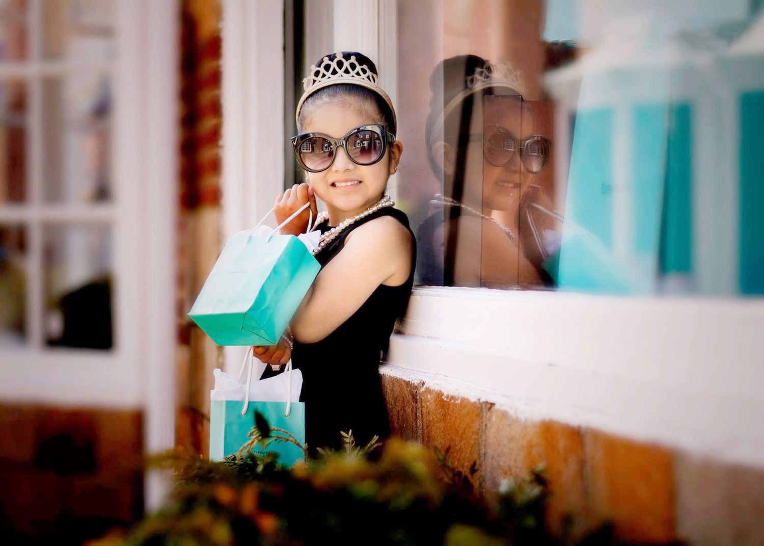 Stylish person with sunglasses and tiara holding shopping bags by storefront window.