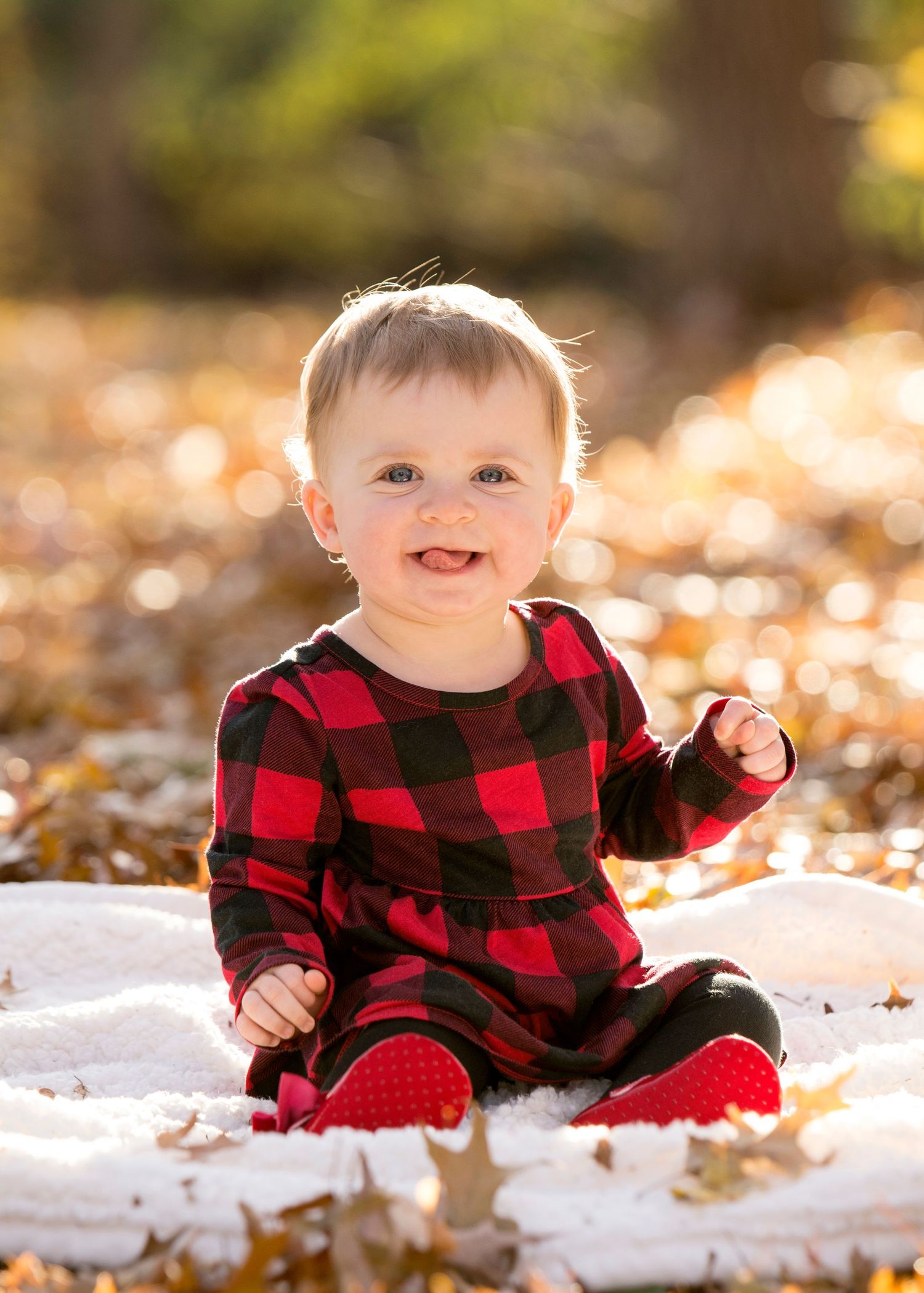 Baby wearing red buffalo plaid outfit sits on white blanket surrounded by fall leaves.