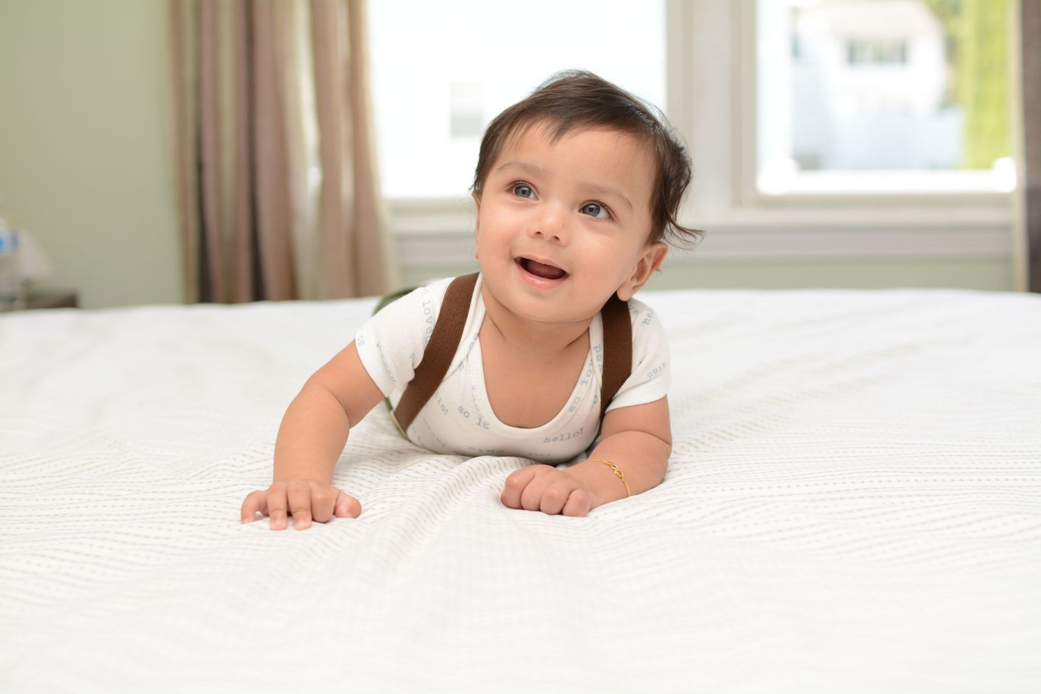 Baby in white onesie crawling on white bedding with curious expression.