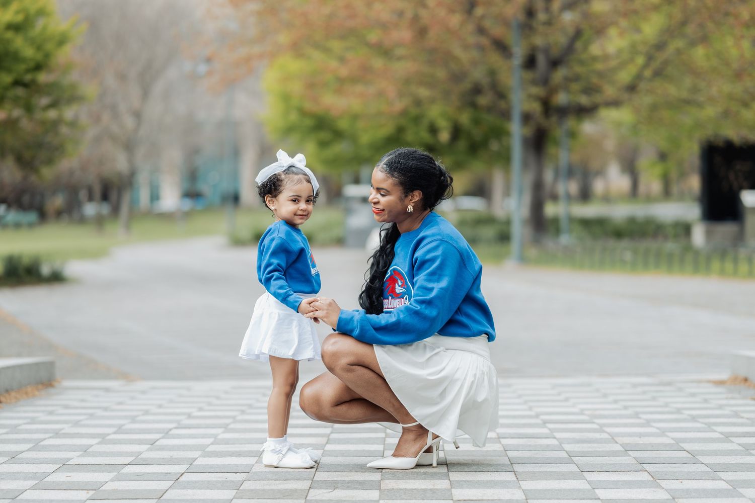 A mother and young daughter in matching blue sweatshirts and white skirts share a joyful moment outdoors.