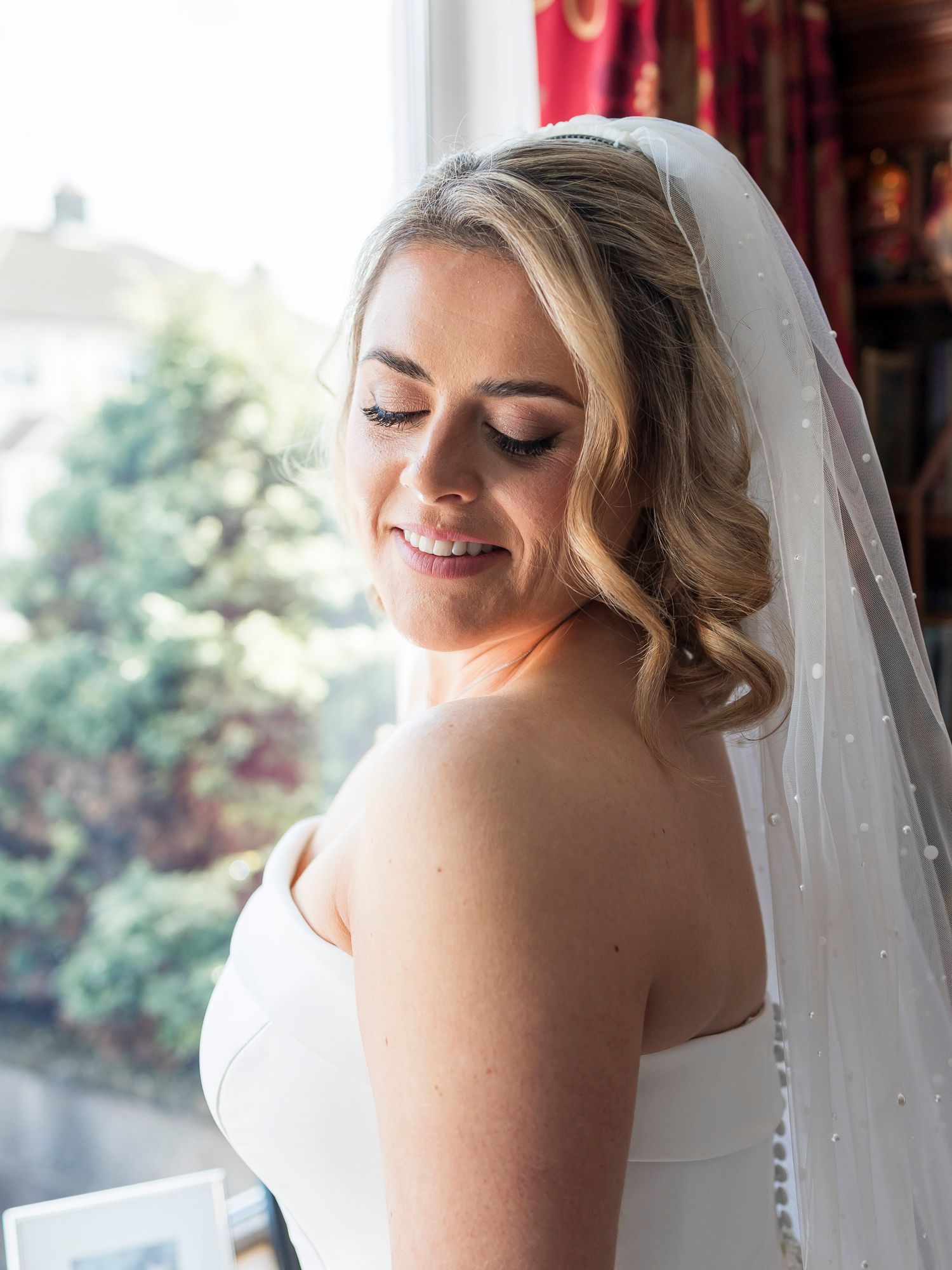 Beautiful bride in white gown and pearl-dotted veil with elegant updo gazing downward by window.
