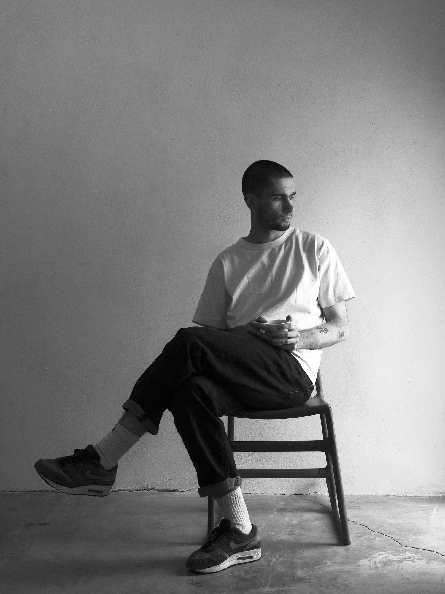 Young man in white t-shirt and dark trousers sitting on wooden chair in minimalist studio.