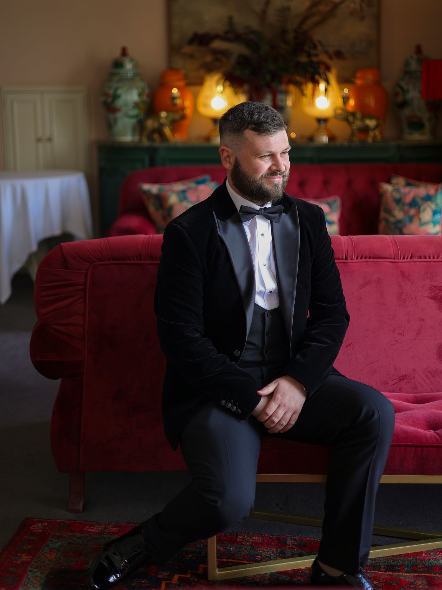 Groom in black velvet tuxedo jacket and bow tie seated on a red velvet sofa at wedding venue.