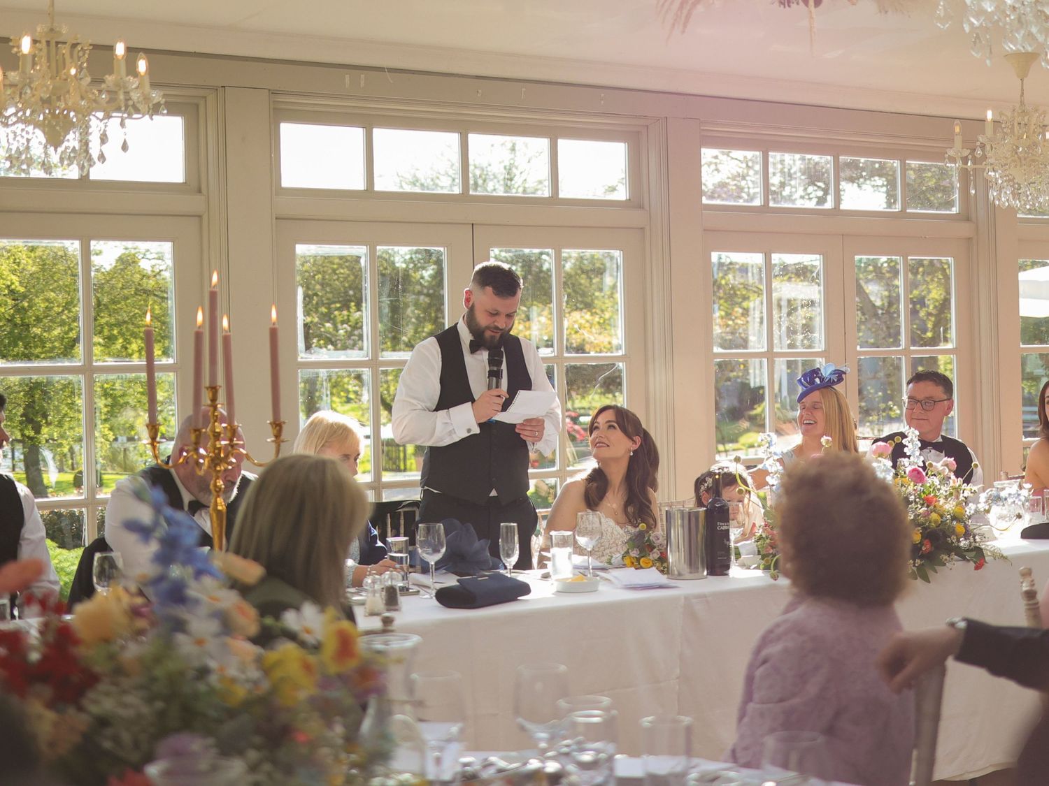 Best man delivering a speech at an elegant wedding reception in a sunlit conservatory venue.