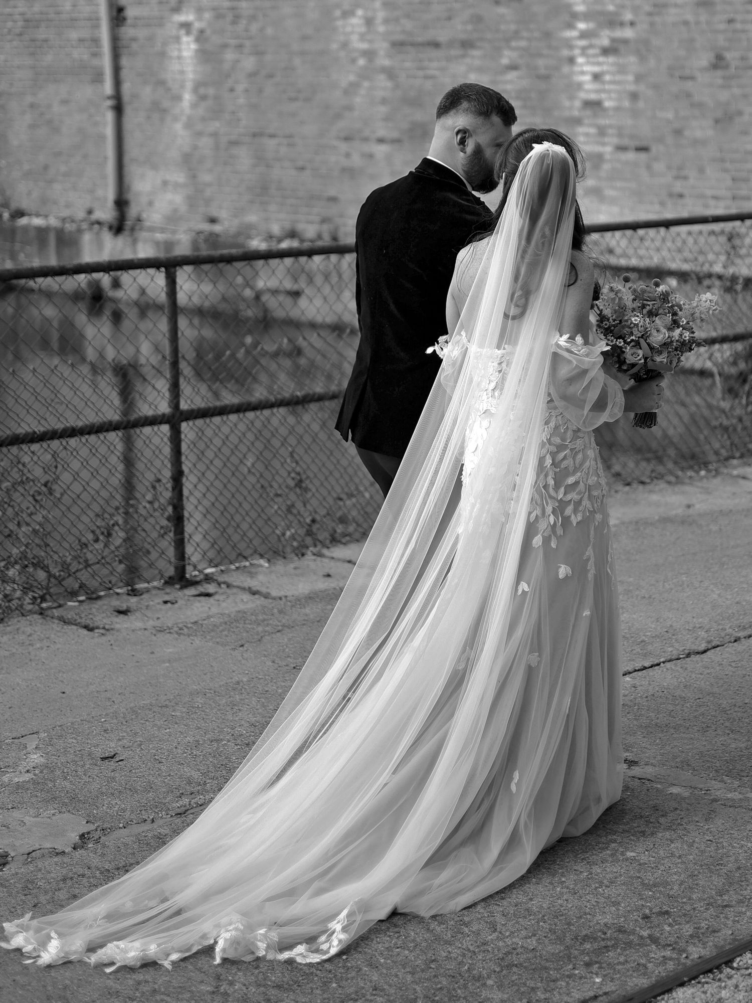 Black and white photo of bride and groom embracing, bride wearing long veil and floral gown, holding bouquet.