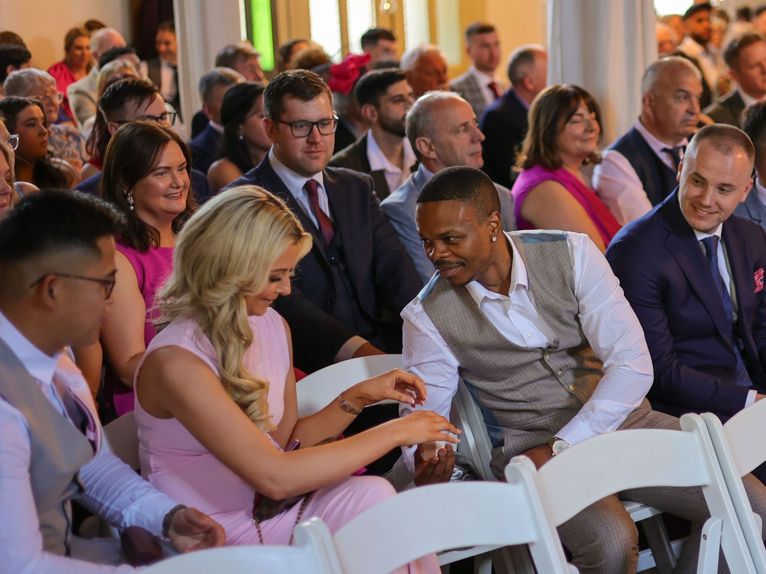 Wedding guests seated in white chairs chatting and smiling during an indoor wedding ceremony.