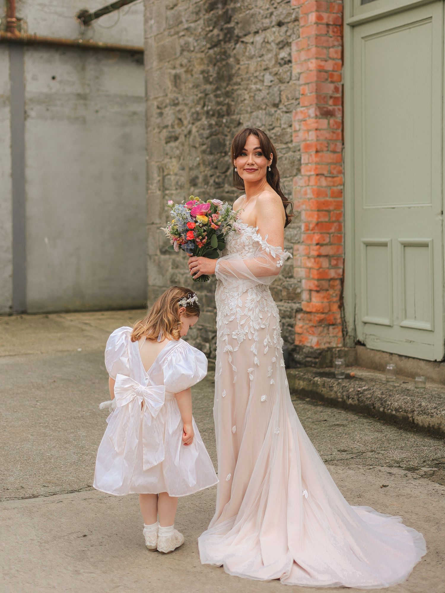 Bride in floral gown holding colorful bouquet stands with flower girl in white dress outside stone building.
