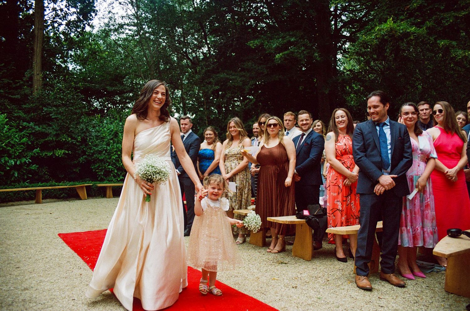 Bride walks red carpet aisle with flower girl at outdoor woodland wedding ceremony with guests.