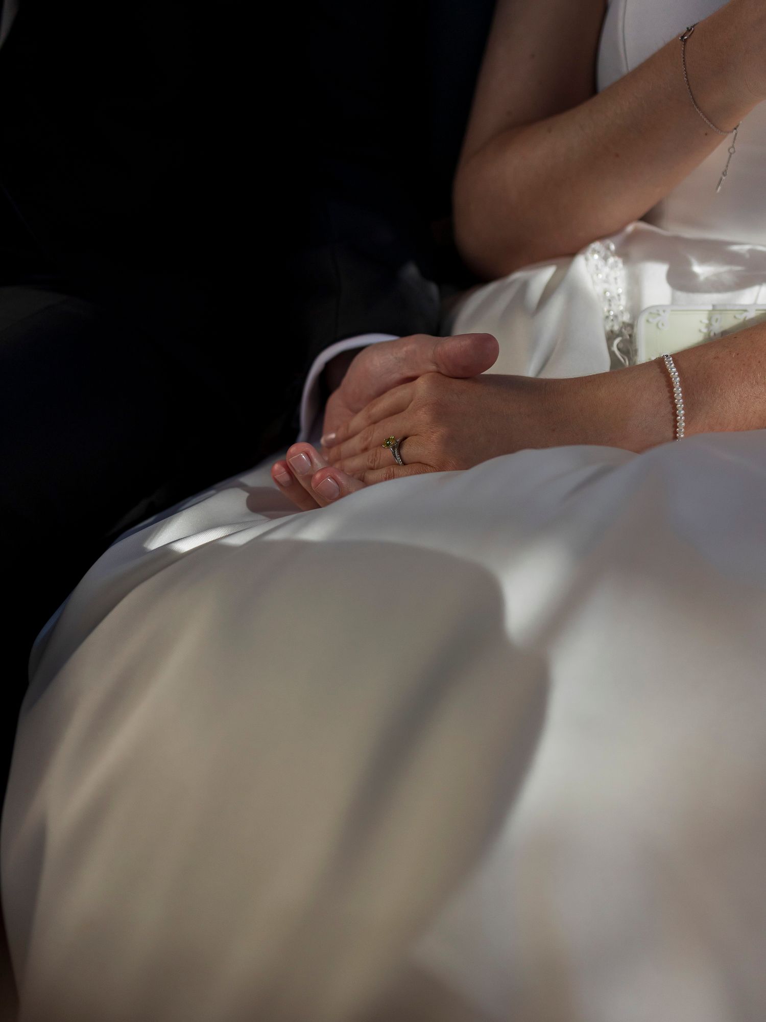 Close-up of bride and groom holding hands, wedding ring visible, in soft natural light.