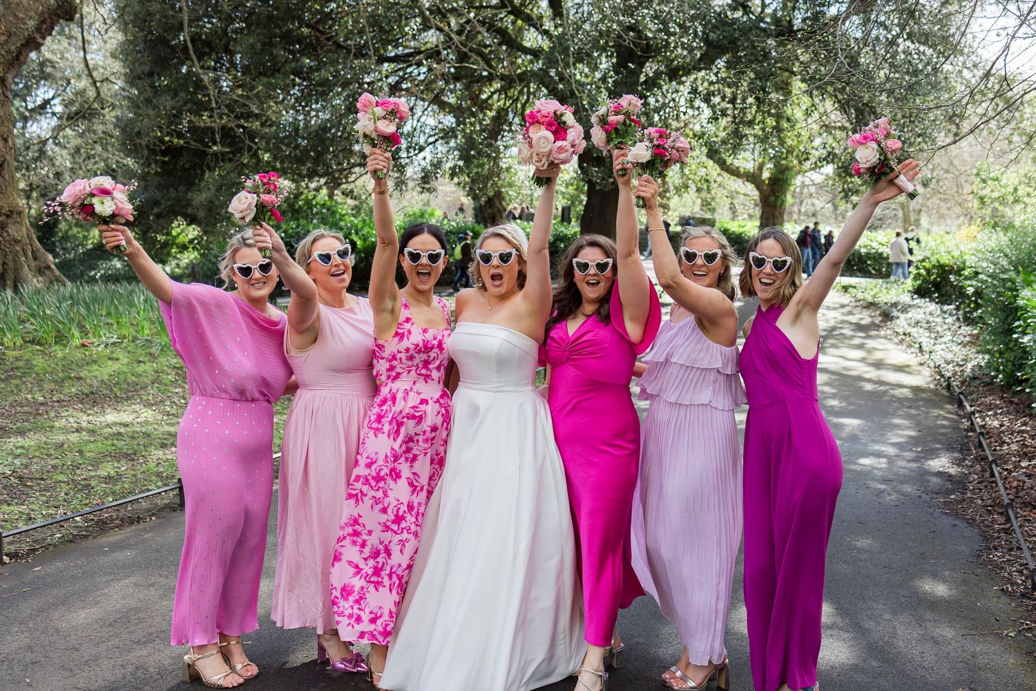 Joyful bride with six bridesmaids in pink dresses raising bouquets outdoors under green trees.