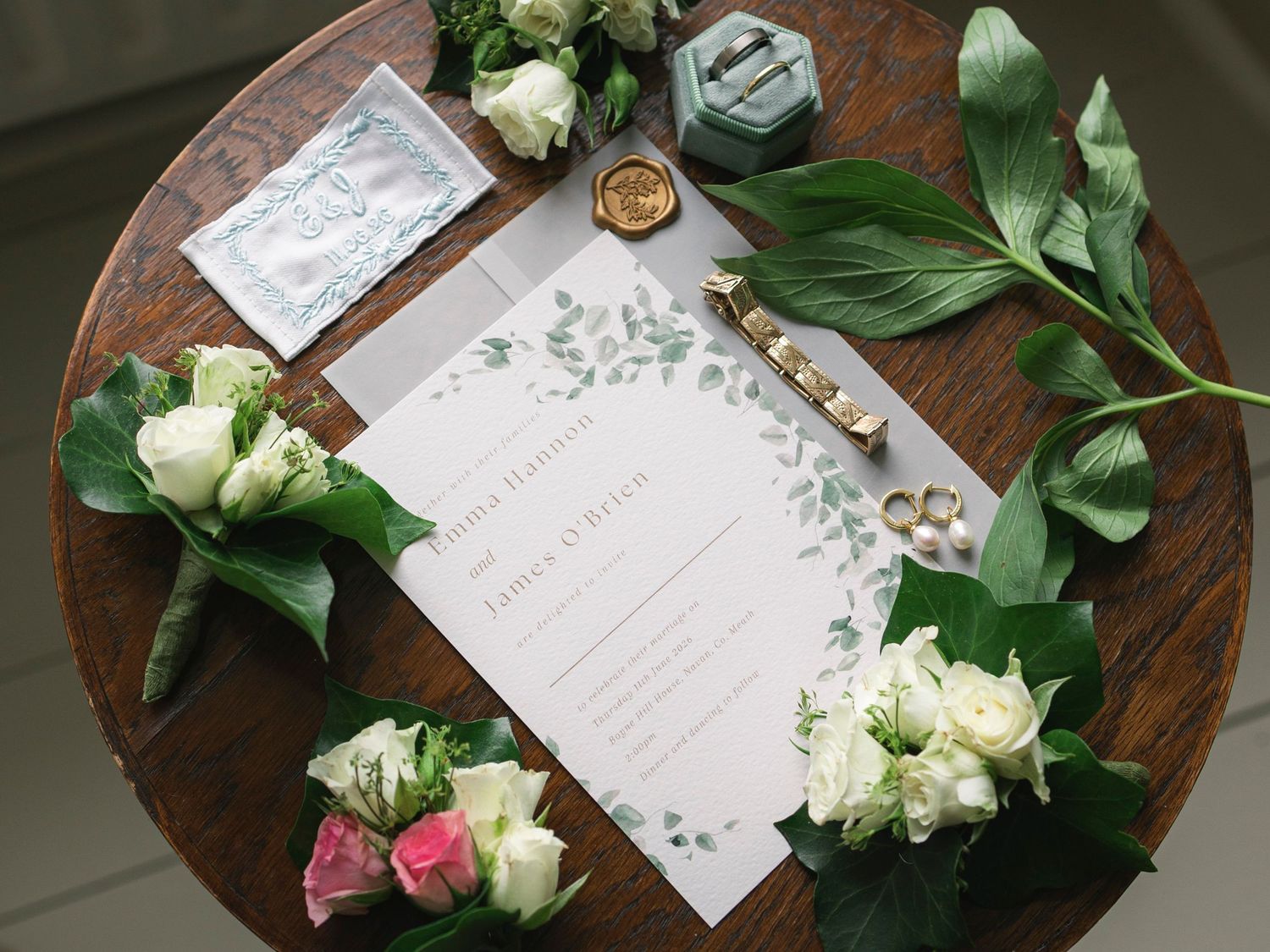 Elegant wedding flat lay with floral invitation, boutonniere, rings, wax seal, and greenery on a wooden table.