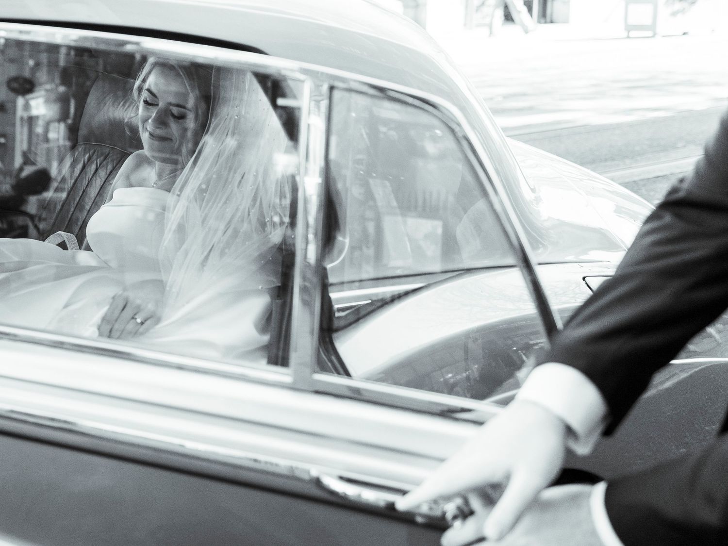 Black and white photo of bride in veil sitting in vintage car as groom opens the classic car door.