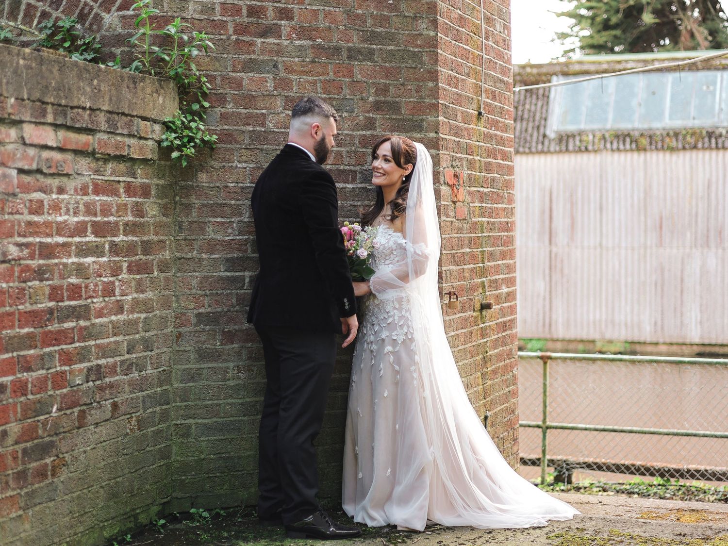 Bride and groom share a romantic moment by a brick wall, bride wearing a white lace gown and veil.