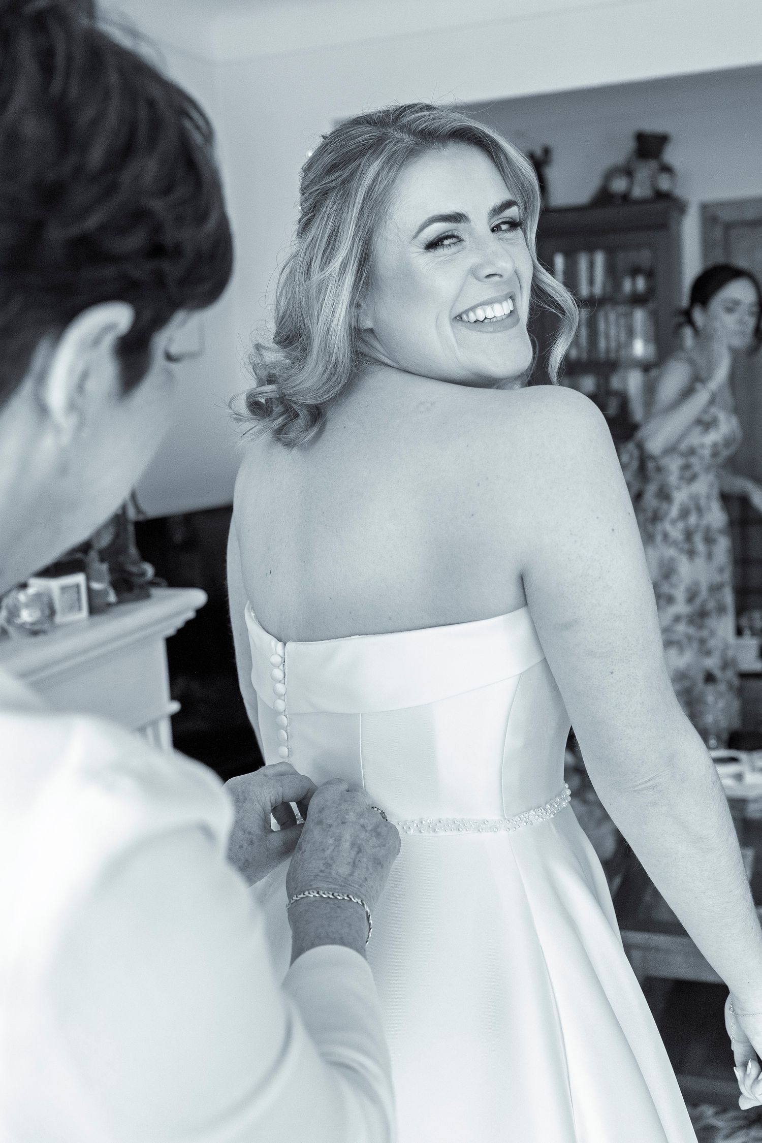 Black and white photo of a smiling bride in a strapless gown having her dress fastened by a woman in a bright room.