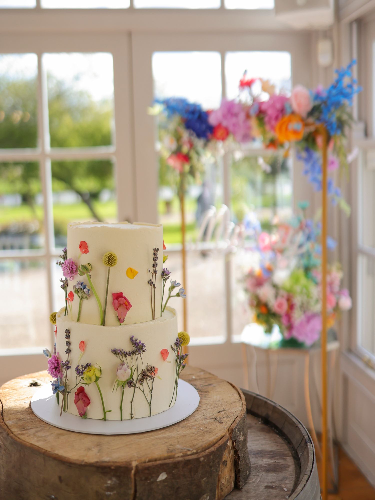 Two-tier white wedding cake decorated with pressed wildflowers displayed on a wooden barrel slice.