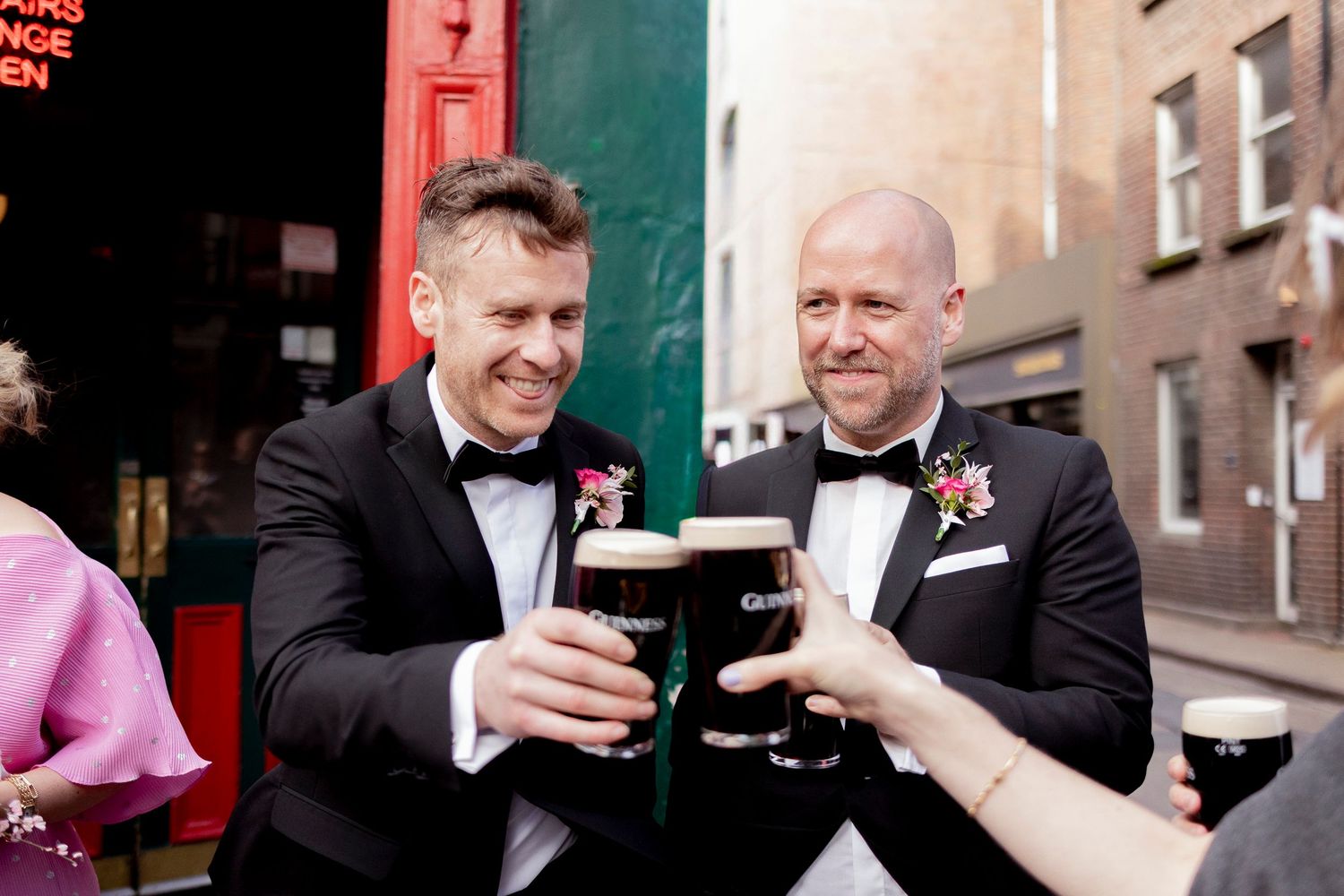 Two groomsmen in tuxedos toast with Guinness pints on city street.