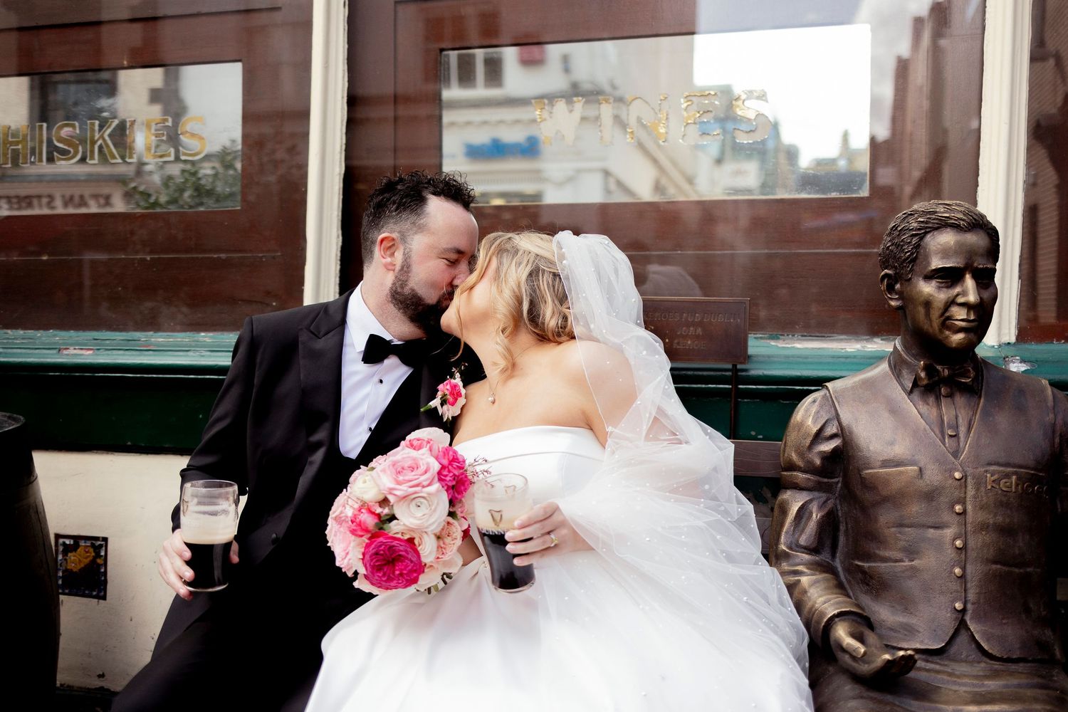 Newlyweds kiss beside bronze statue on Dublin street holding pink bouquet.