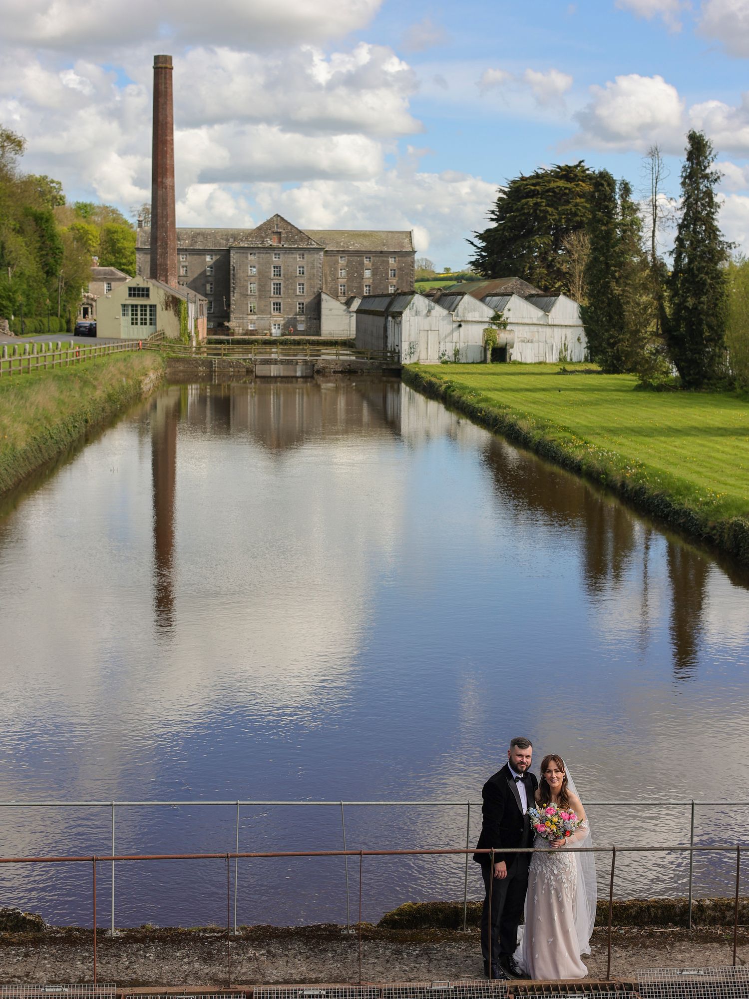 Bride and groom pose on a bridge over a calm canal with a historic mill building reflected in the water.