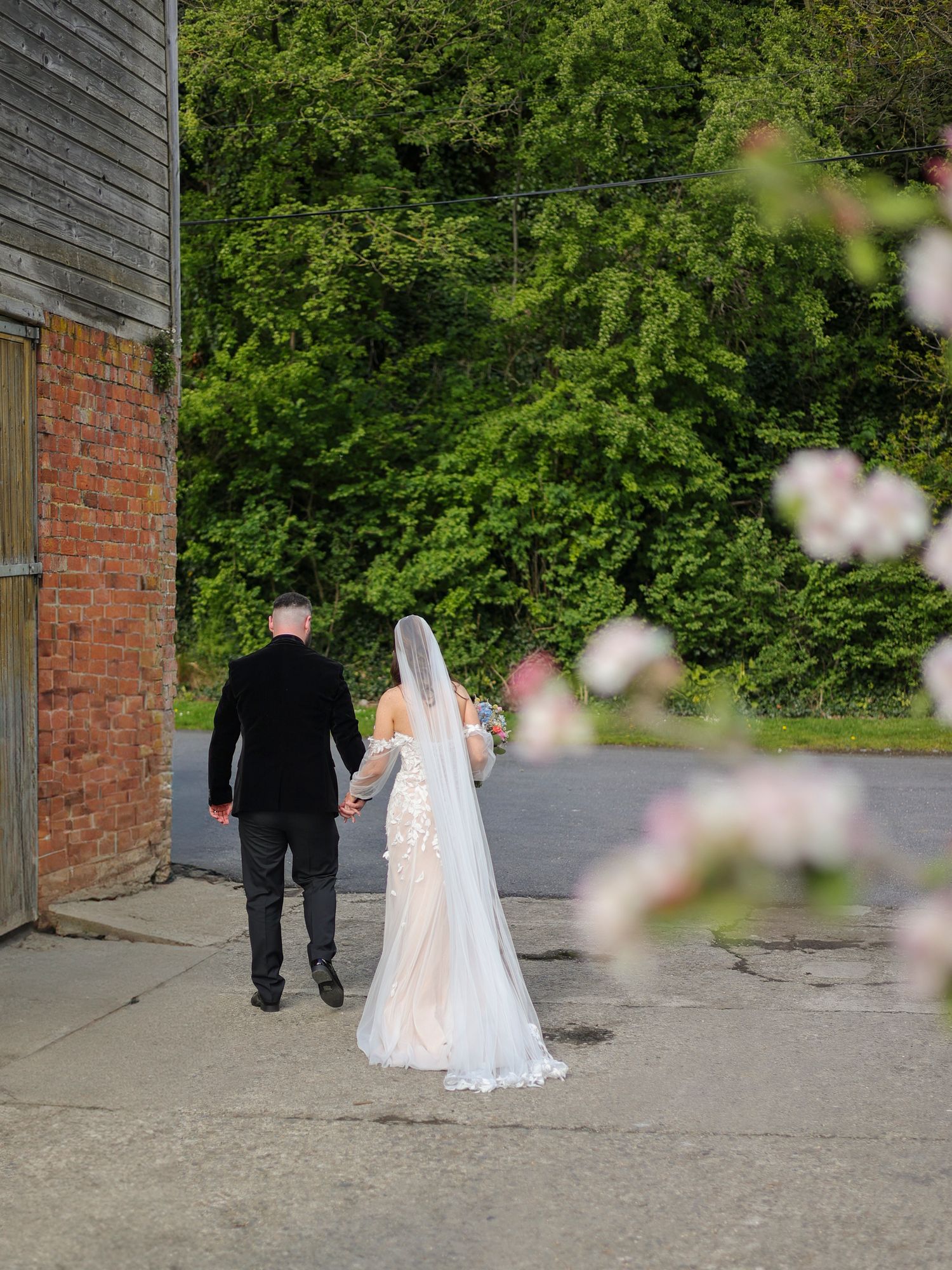 A bride in a white gown and veil walks hand-in-hand with a groom past a rustic brick barn surrounded by lush greenery.