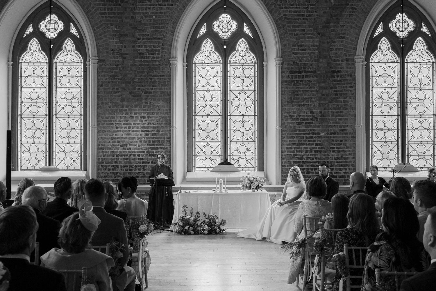Black and white wedding ceremony photo in Gothic church with arched windows and guests seated in pews.