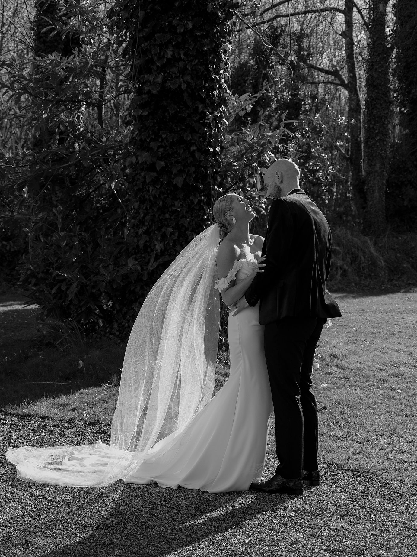Bride and groom standing together in a garden, bride wearing a long flowing veil, black and white photo.