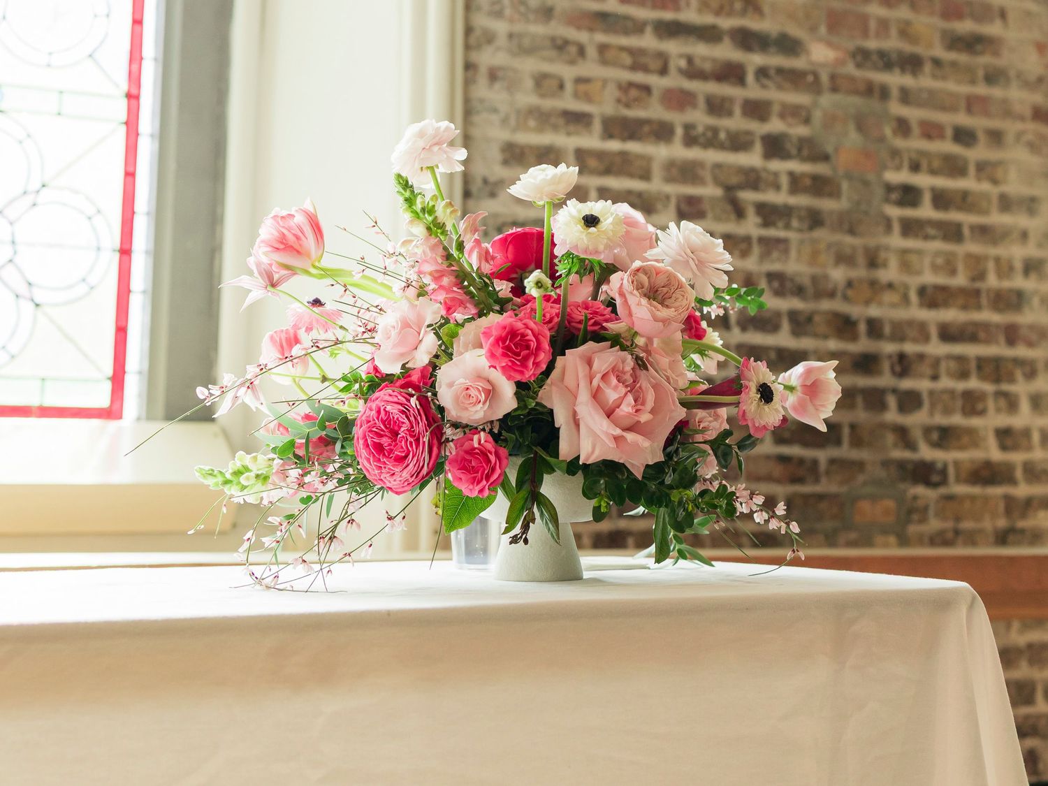 Pink roses and white ranunculus floral arrangement in white vase on table near brick wall window.