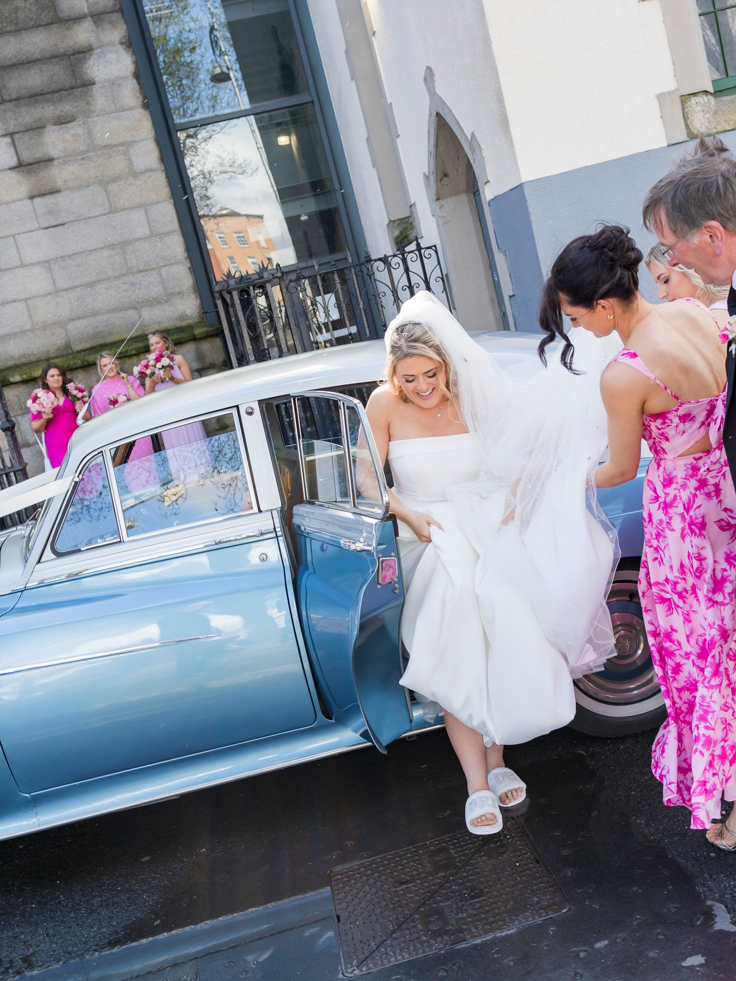 Bride in white strapless gown and veil exits a vintage blue car assisted by guests in pink dresses.