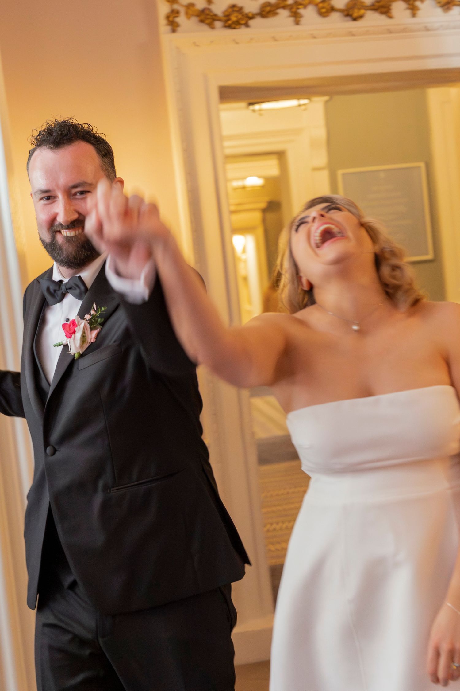 Joyful bride and groom make grand entrance at wedding reception, laughing and holding hands in elegant venue.