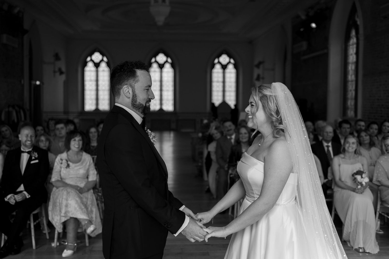 Black and white wedding ceremony photo of bride and groom holding hands at the altar in a church.