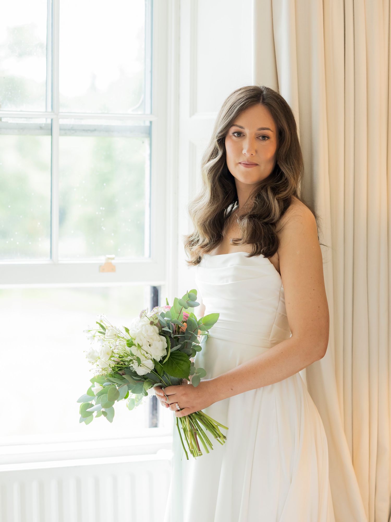 Elegant bride in white strapless gown holding white and green bouquet, standing by window in soft natural light.